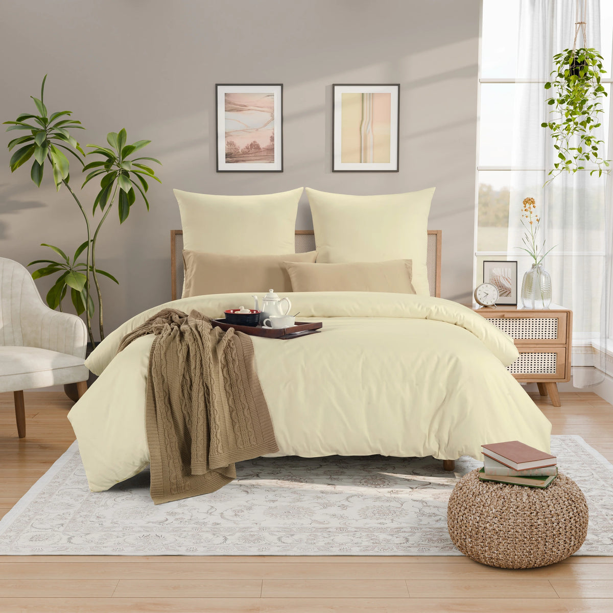 A neatly made bed in a bright bedroom setting, dressed in a pale buttery yellow duvet cover and matching pillow shams, with coordinating beige pillows and a taupe throw blanket draped across the foot of the bed. The room features a light gray wall, two framed abstract prints, a white armchair, a wooden nightstand, and a woven pouf.