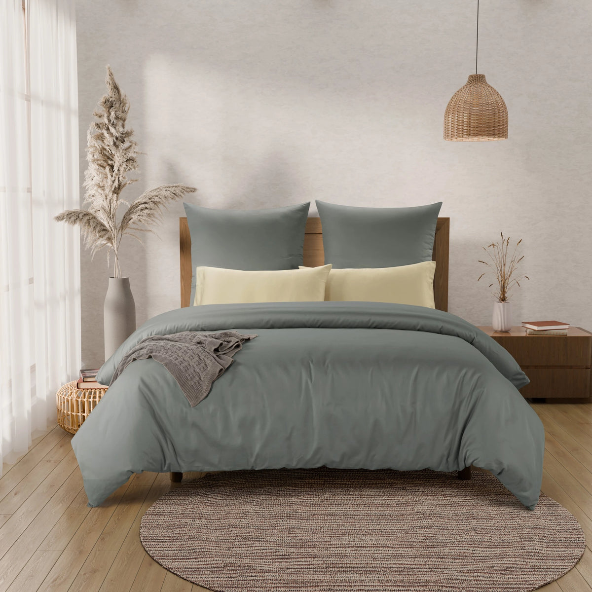 A styled bedroom scene showing a bed made up with a muted sage green duvet cover and matching pillow shams, plus two pale yellow pillows. The room has a textured light wall, wood floor, pendant light, side table, and decorative dried grasses in vases.