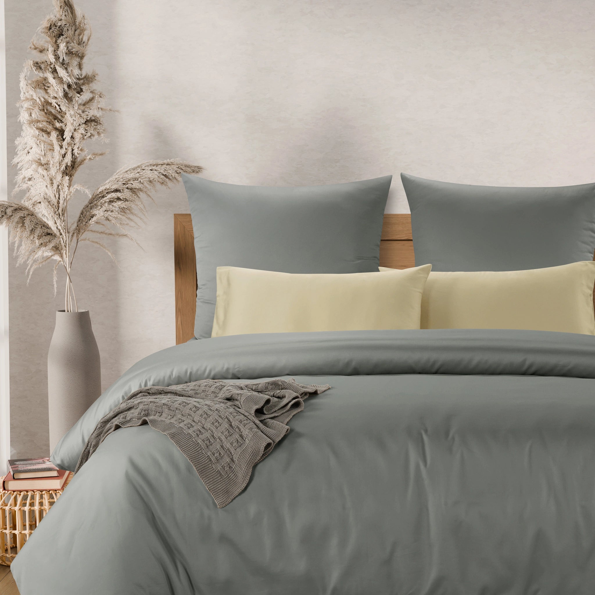 A neatly made bed with a muted sage green duvet cover and matching pillow shams, styled with two pale yellow accent pillows, a wooden headboard, a draped gray throw blanket, and a vase of dried grass on a side table.