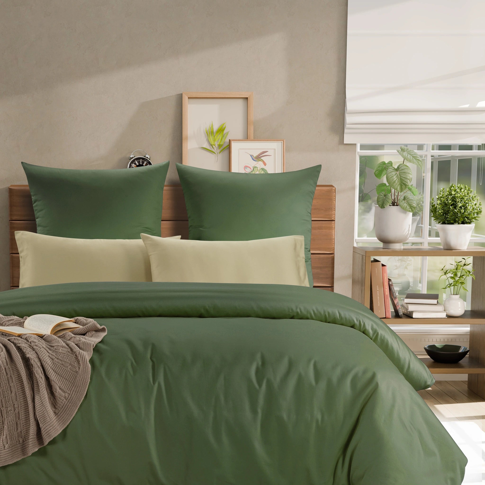 A styled bed in a bedroom setting with an olive green duvet cover and matching pillow shams, layered with two cream-colored accent pillows. The room features a wooden headboard, a window with a white roller blind, and a small wooden side table decorated with potted plants and books.